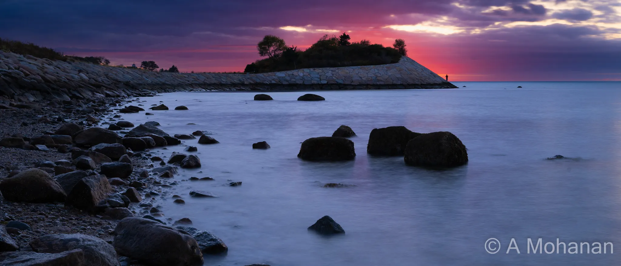 Long-exposure seascape at sunset, silky water around dark rocks, stone causeway leading to a lone silhouetted figure, dramatic purple and crimson sky.