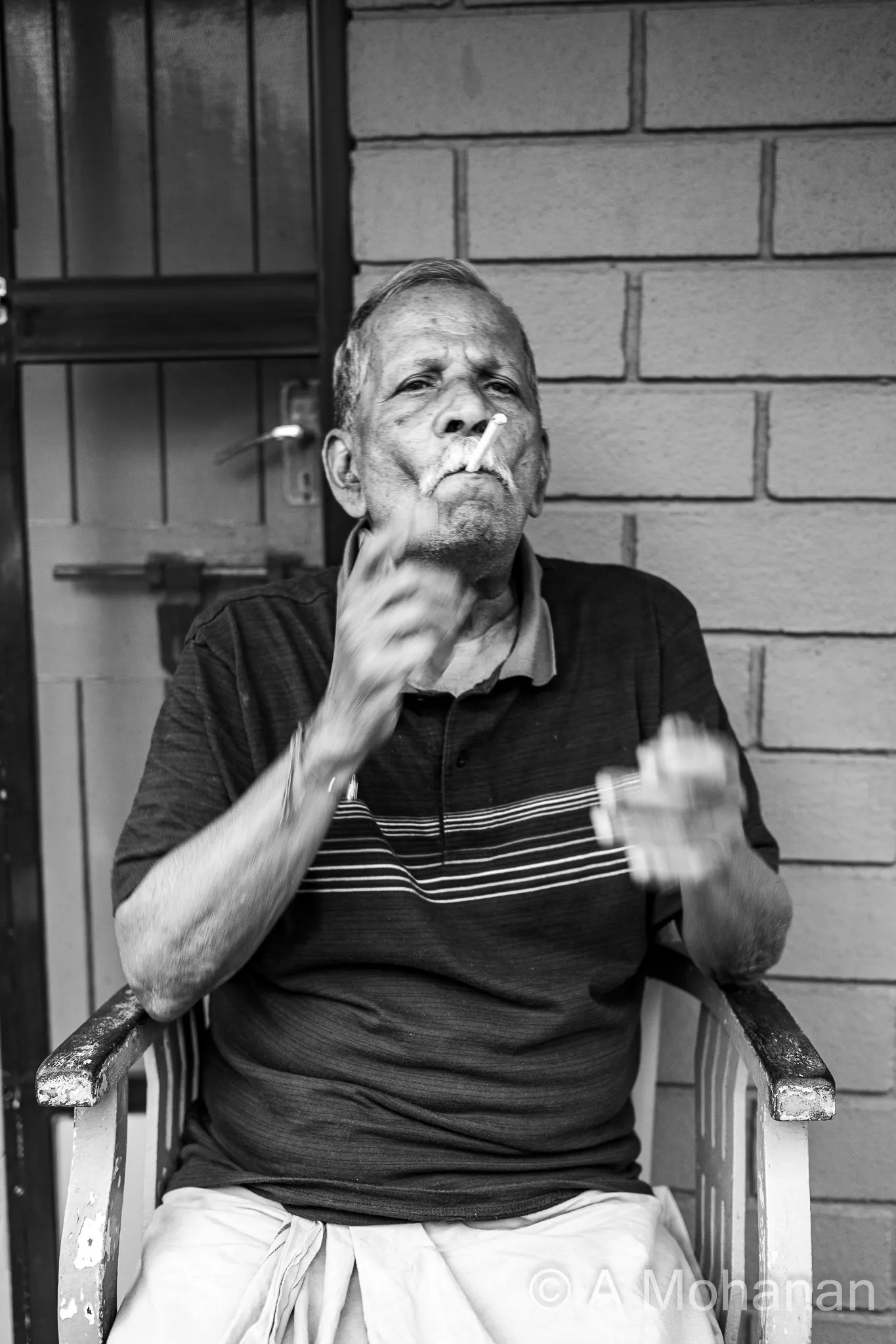 An elderly man mid-gesture, motion-blurred hands, cigarette, seated on a verandah against a brick wall, black and white.