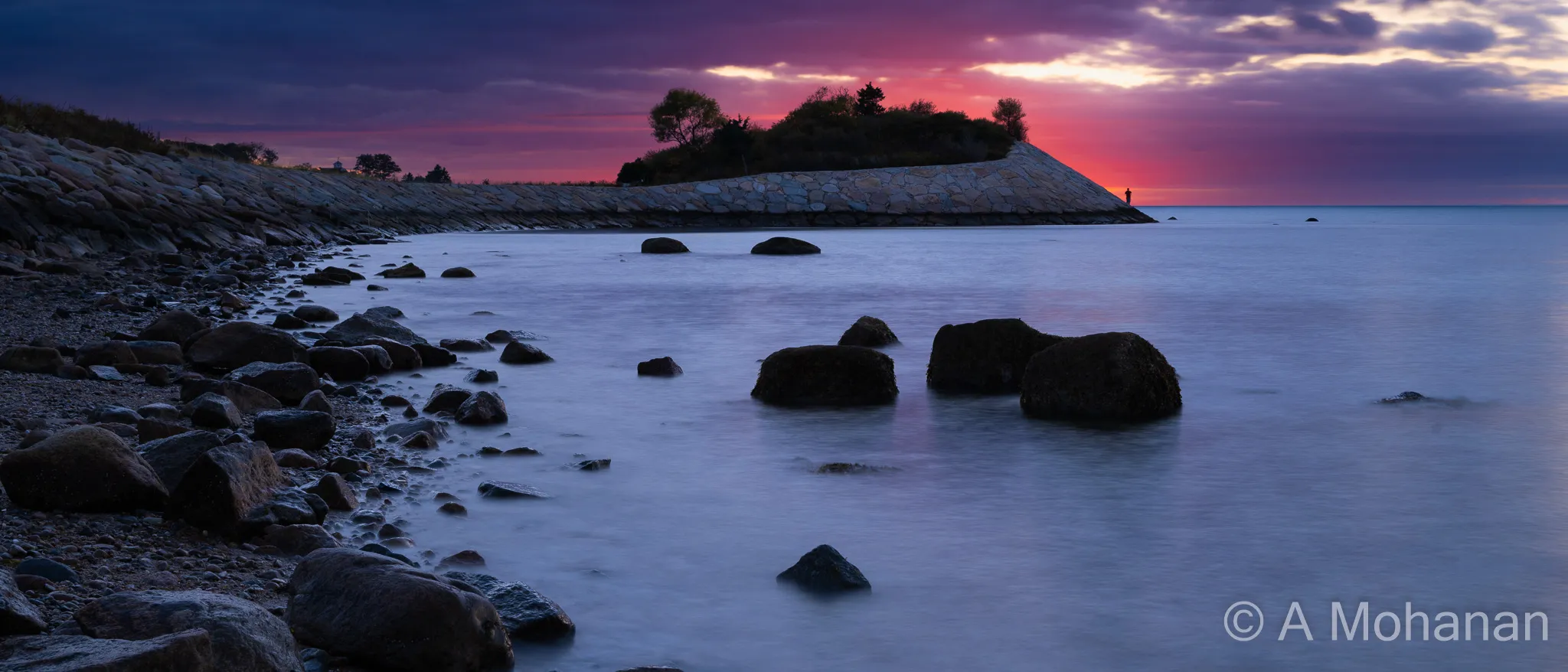 Long-exposure seascape at sunset, silky water around dark rocks, stone causeway leading to a lone silhouetted figure, dramatic purple and crimson sky.