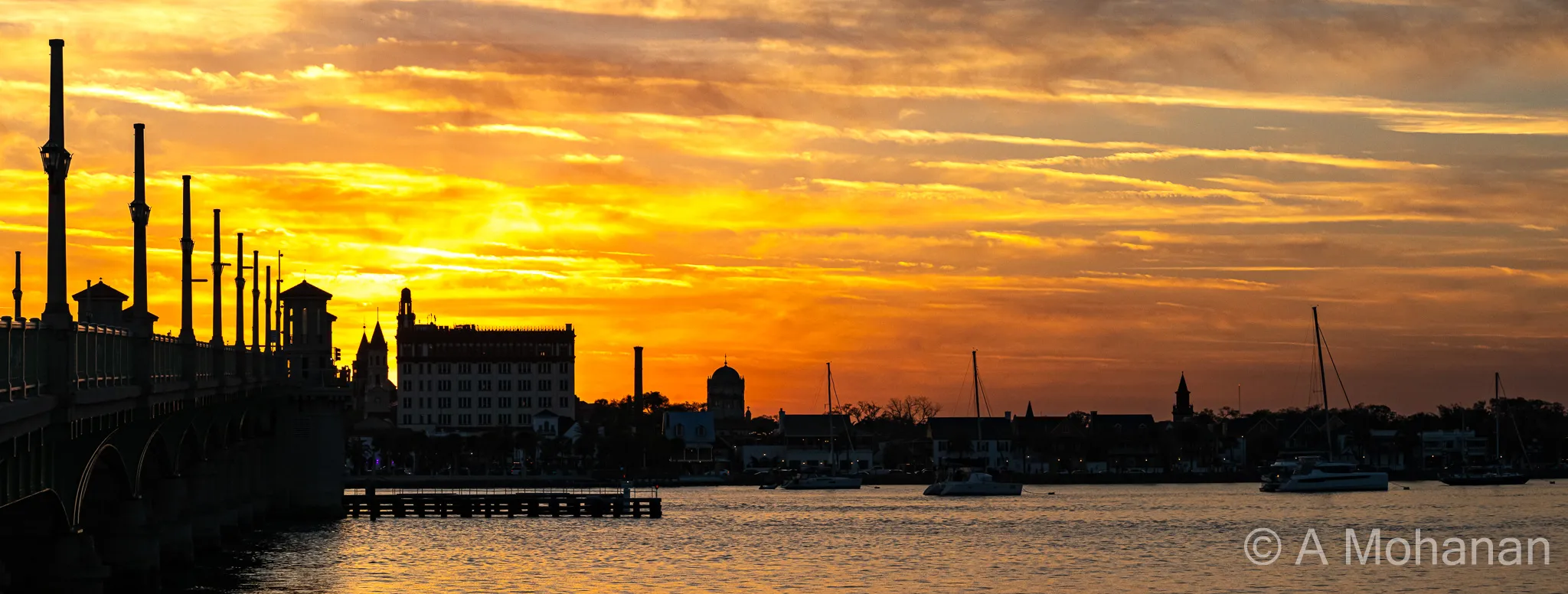 St. Augustine skyline silhouetted against a blazing orange sunset sky, Bridge of Lions in the foreground, sailboats on the Matanzas River.