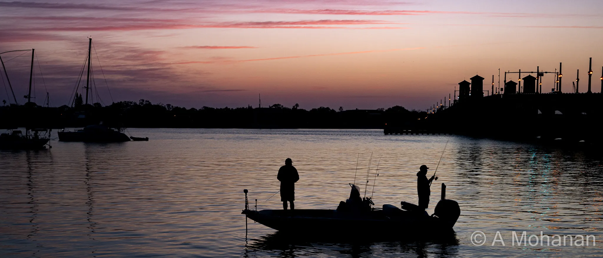 Two fishermen silhouetted on a boat in pre-dawn stillness, mauve and salmon sky reflected in calm water, Bridge of Lions visible in the distance.
