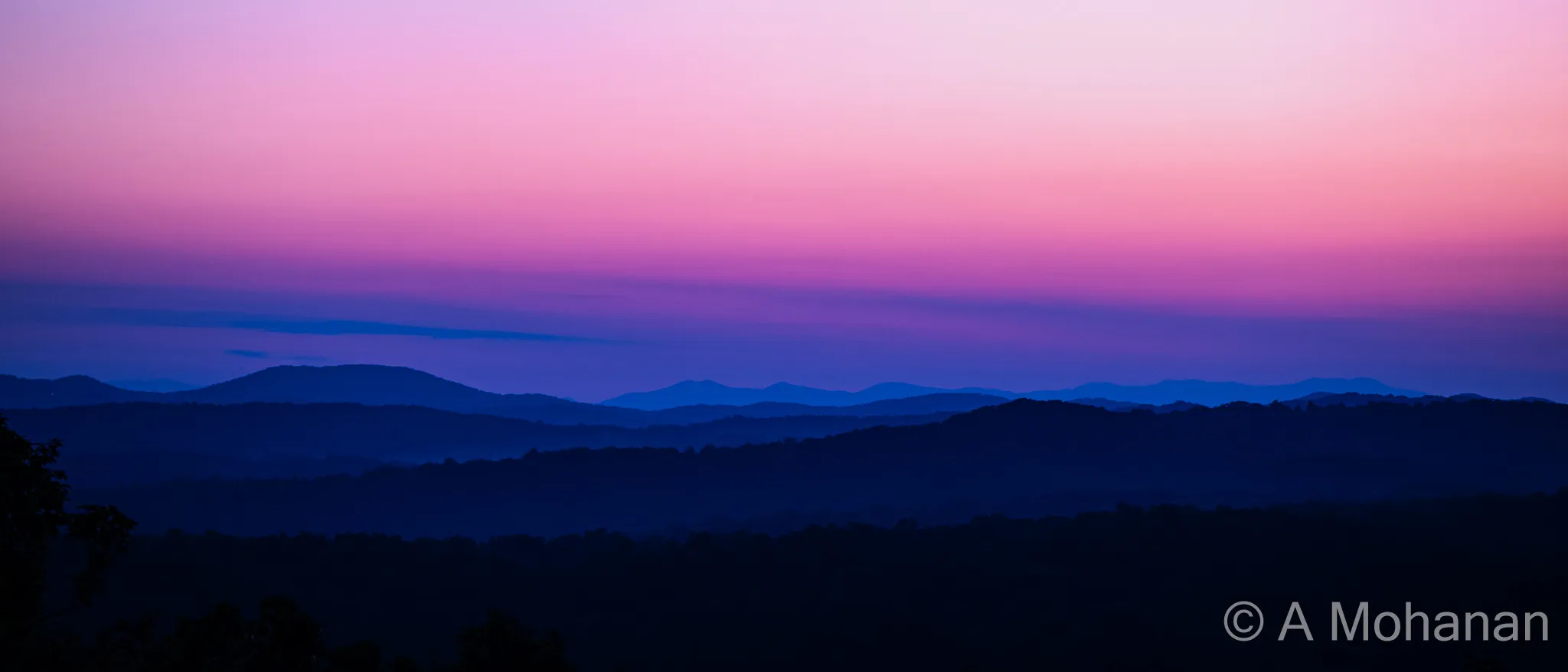 Layered mountain ridgelines silhouetted in blue haze beneath a twilight sky of violet, magenta and salmon.