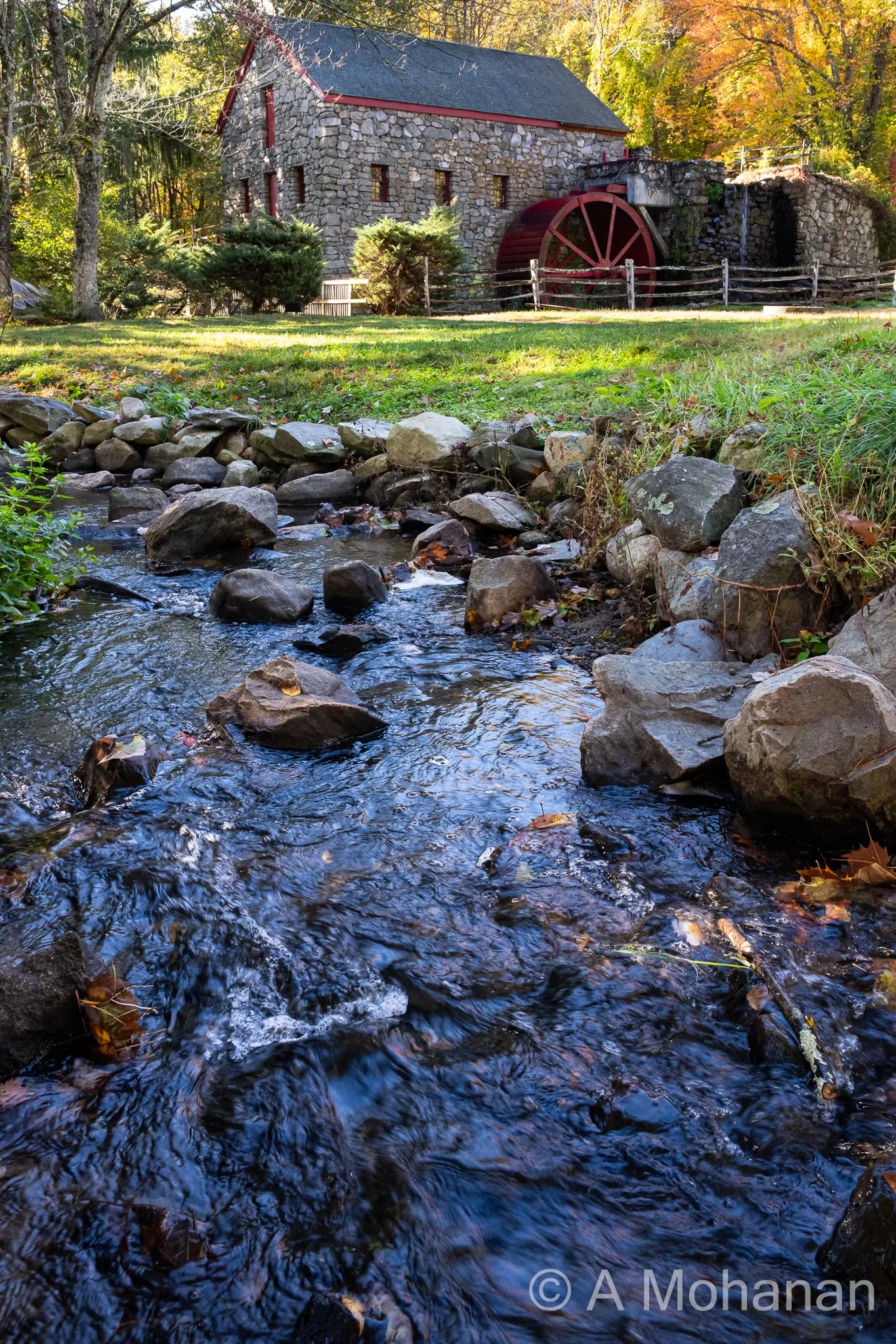 Rocky stream leading to a historic stone grist mill with a red waterwheel, surrounded by early autumn foliage.